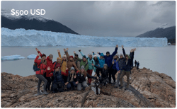 TrovaTrip group of travelers wearing rainbow coats smiling with hands in air in front of glacier