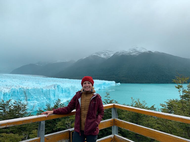 TrovaTrip traveler in red hat smiling in front of glacier with snow mountains behind her