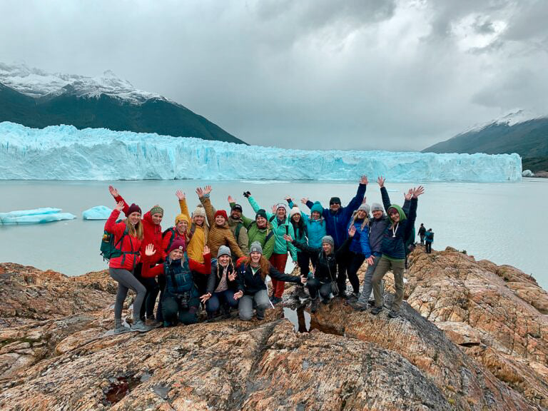 TrovaTrip group of travelers in rainbow ski jackets smiling with hands in the air in front of a glacier and mountain behind them