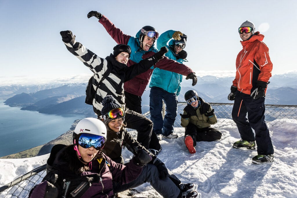 TrovaTrip group of travelers in snowboarding gear smiling and posing on top of a snowy mountain with snow and lake behind them in New Zealand