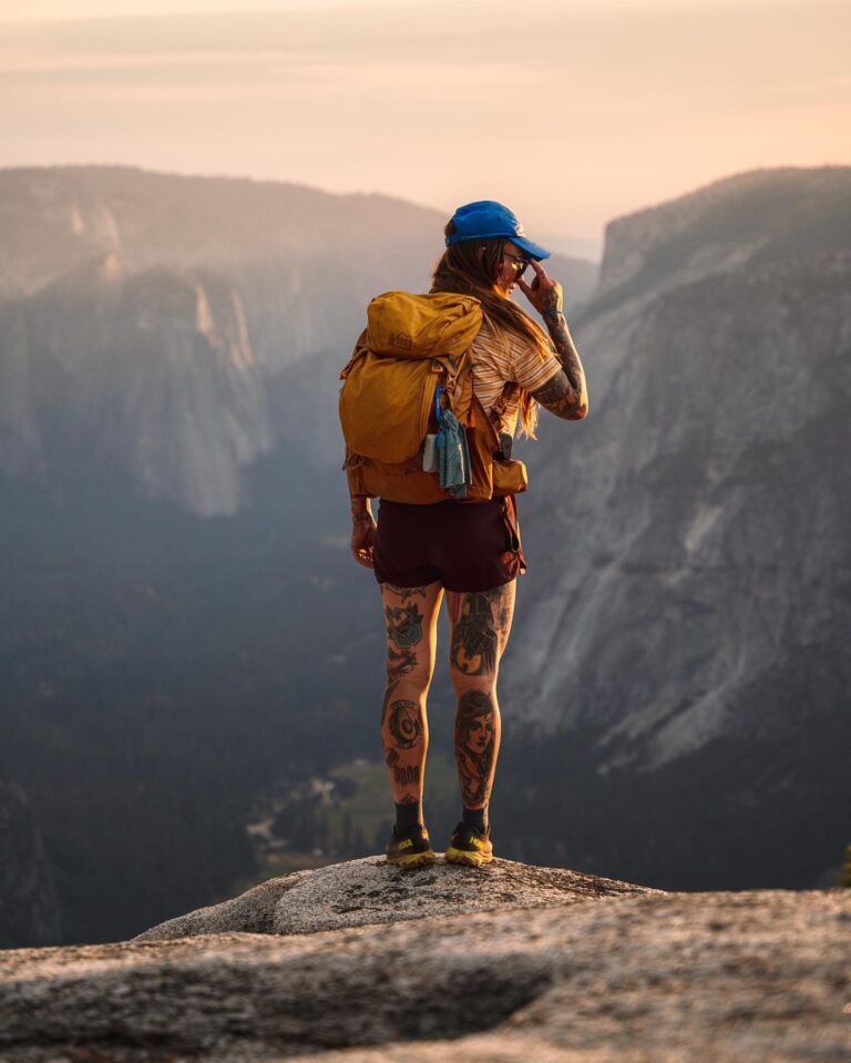 TrovaTrip woman with tattoos standing on edge of mountain with yellow backpack