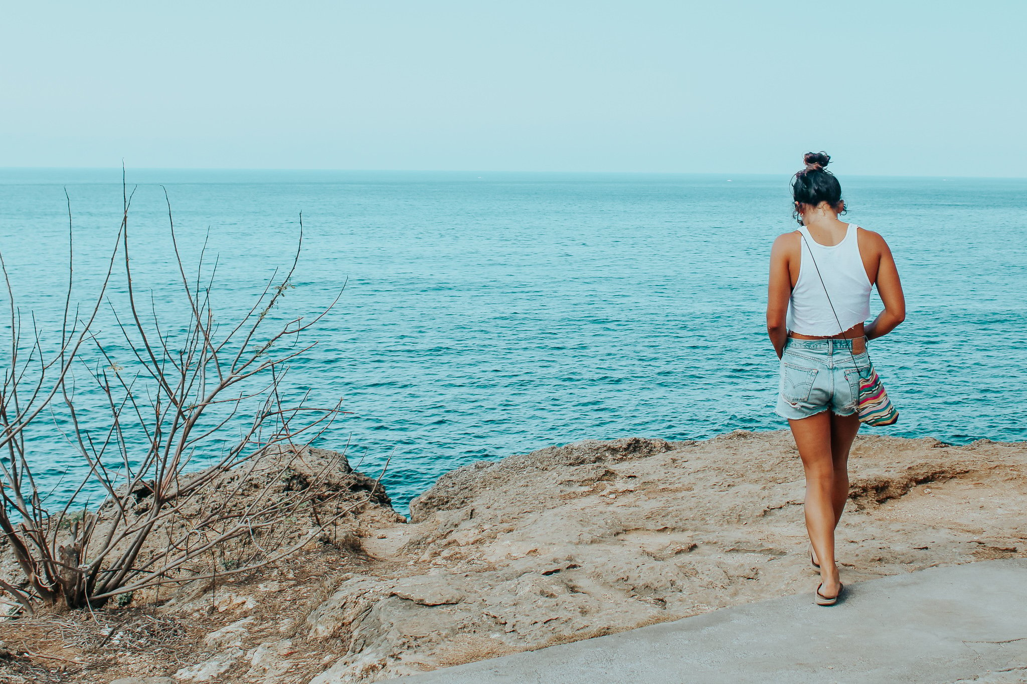 TrovaTrip woman in white shirt and blue shorts walking away from camera towards sand overlooking clear blue ocean water