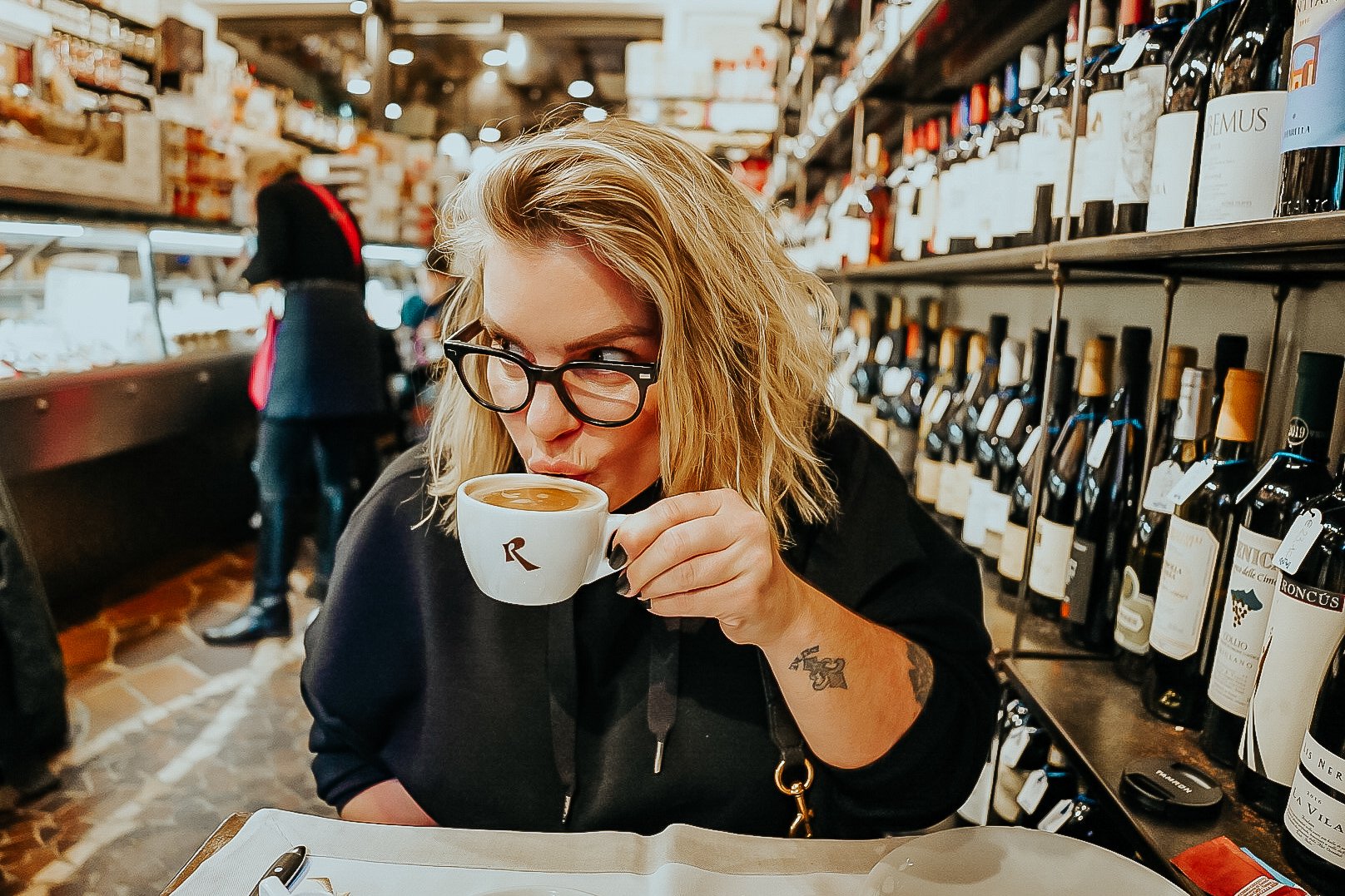 TrovaTrip woman in glasses sipping an espresso in a cafe with wine bottles behind her in Italy