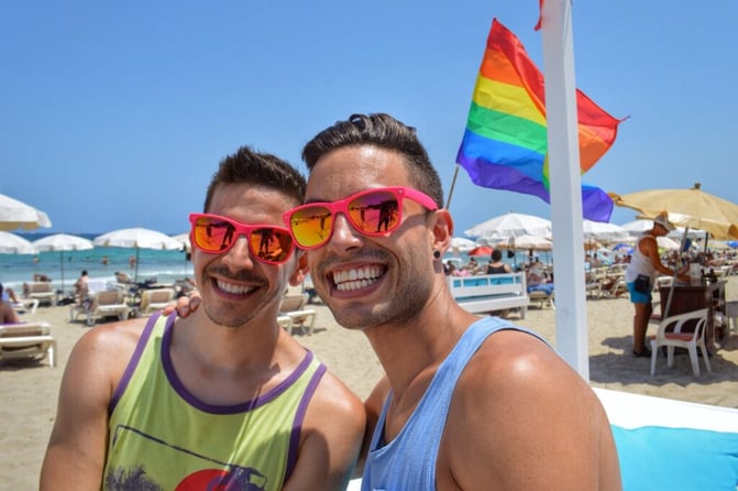 TrovaTrip two men with pink sunglasses smiling on the beach with ocean and pride flag behind them