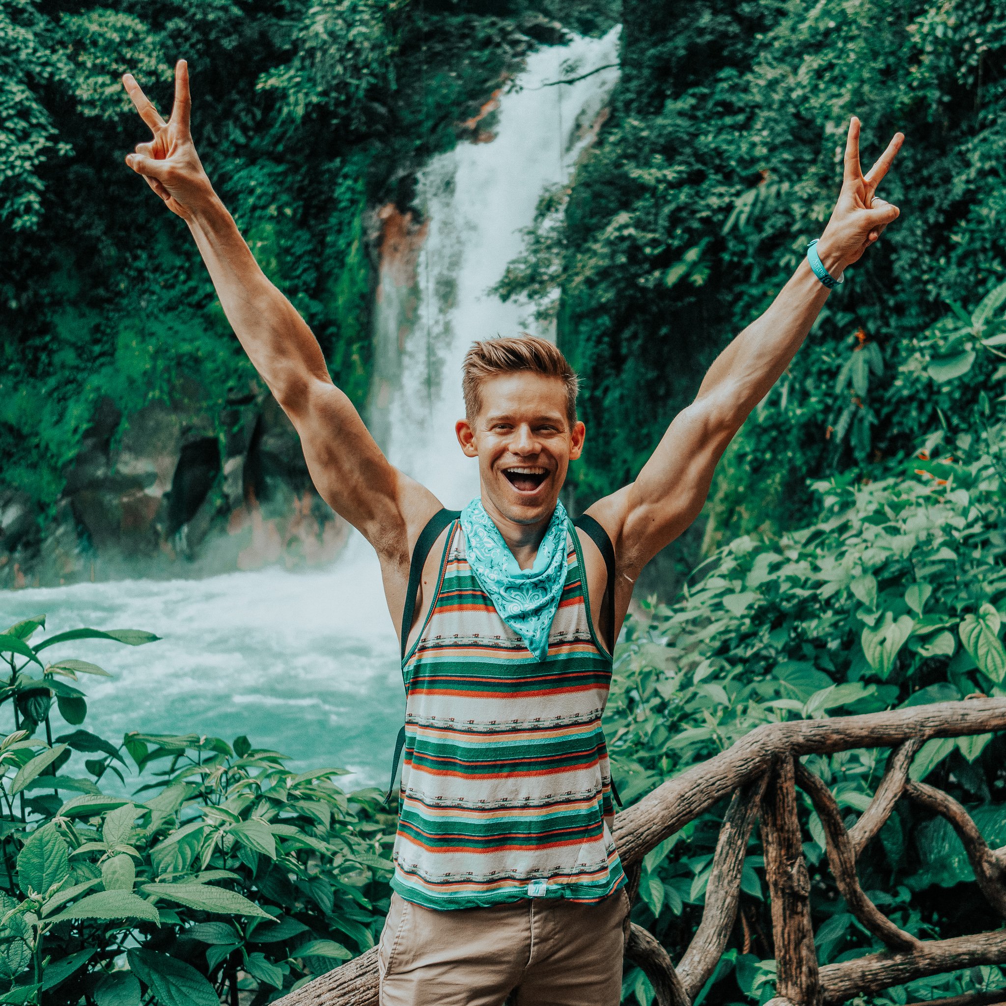 TrovaTrip man holding up peace signs with both arms in the air in front of a lush green waterfall in the Costa Rica jungle