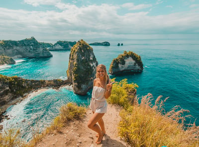 TrovaTrip Bali Nusa Penida woman standing on ledge facing camera and pointing to ocean behind her
