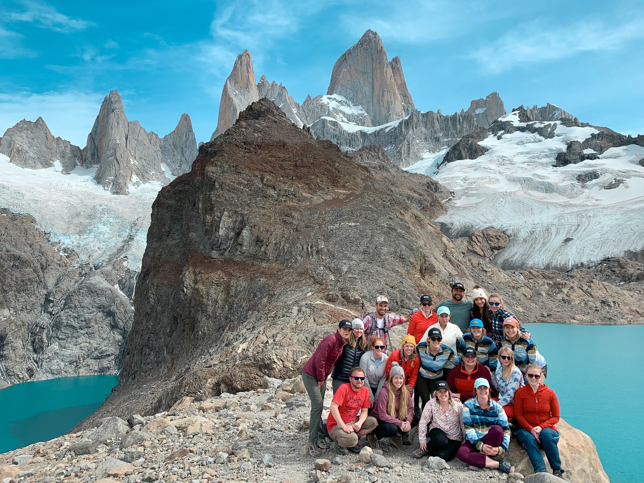 TrovaTrip group of travelers smiling in front of large rock formation with clear blue water below them and snowy mountains behind them in Patagonia