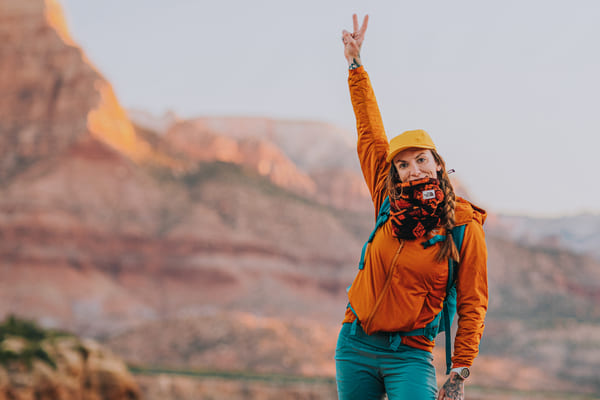 TrovaTrip woman in orange jacket holding up peace sign with red mountains behind her in Zion