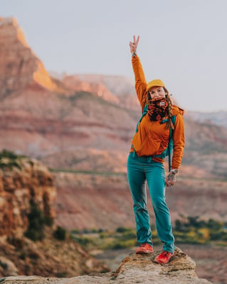 TrovaTrip Zion woman in orange jacket holding peace sign up with canyons behind her